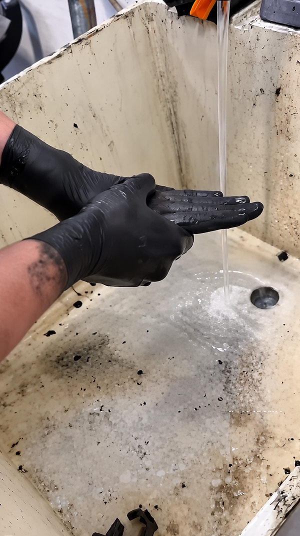 Mechanic's hands coated in thick black grease held under a running faucet at a shop sink, before washing with Grip Clean hand soap